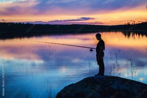 Темный мужской силуэт рыбака с удочкой, стоящего на большом камне на фоне неба и заката, отражающегося в воде фотография Stock